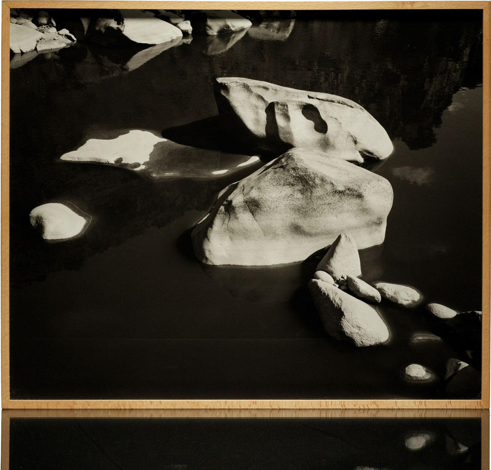 A framed black-and-white print: pale boulders in dark, still water, in a light wood frame