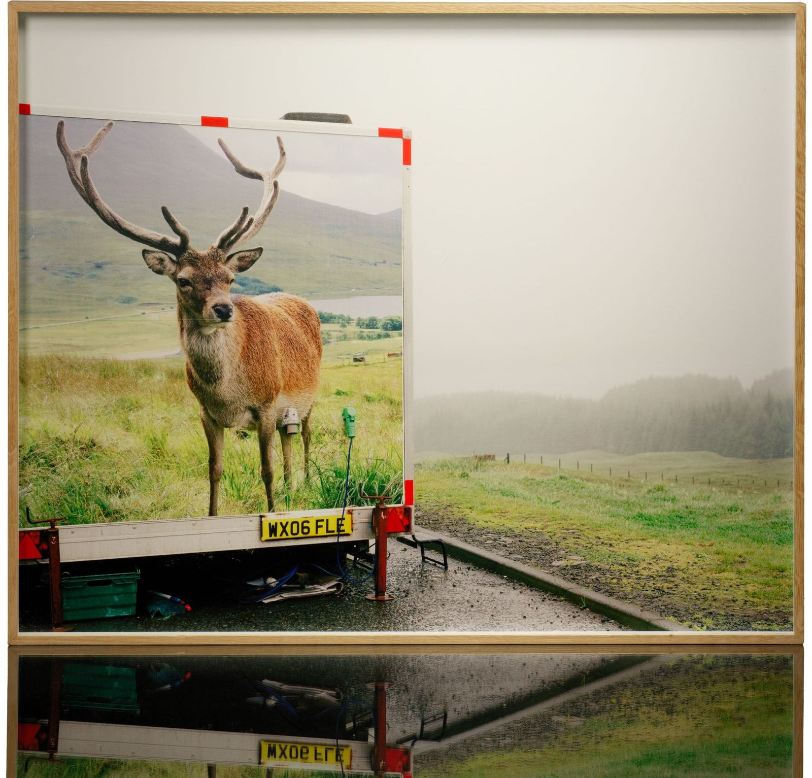 A framed colour print: a trailer printed with a picture of a stag, parked by a road on a misty moor, in a light wood frame