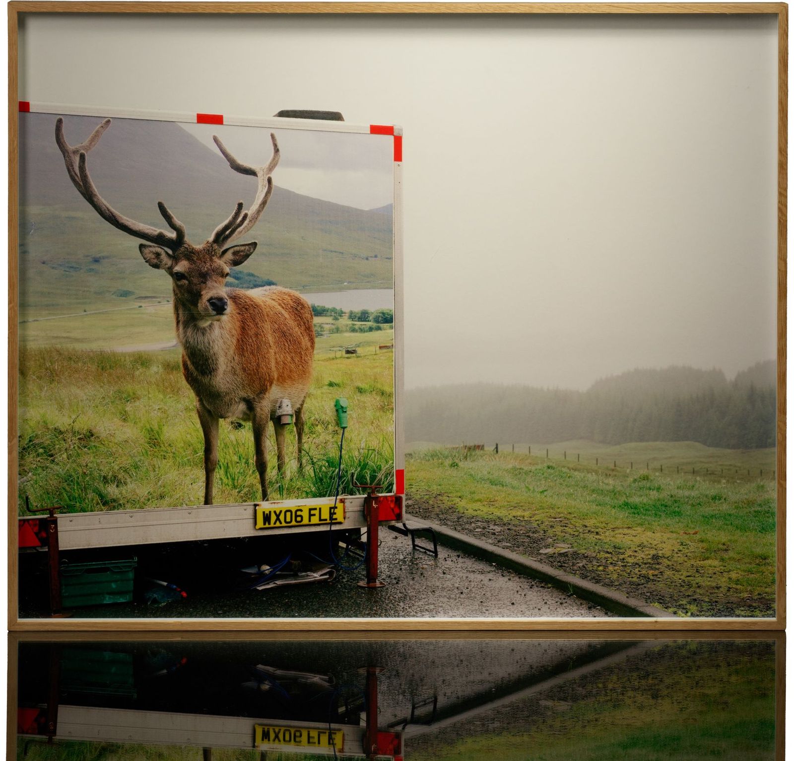 A framed colour print: a trailer printed with a picture of a stag, parked by a road on a misty moor, in a light wood frame