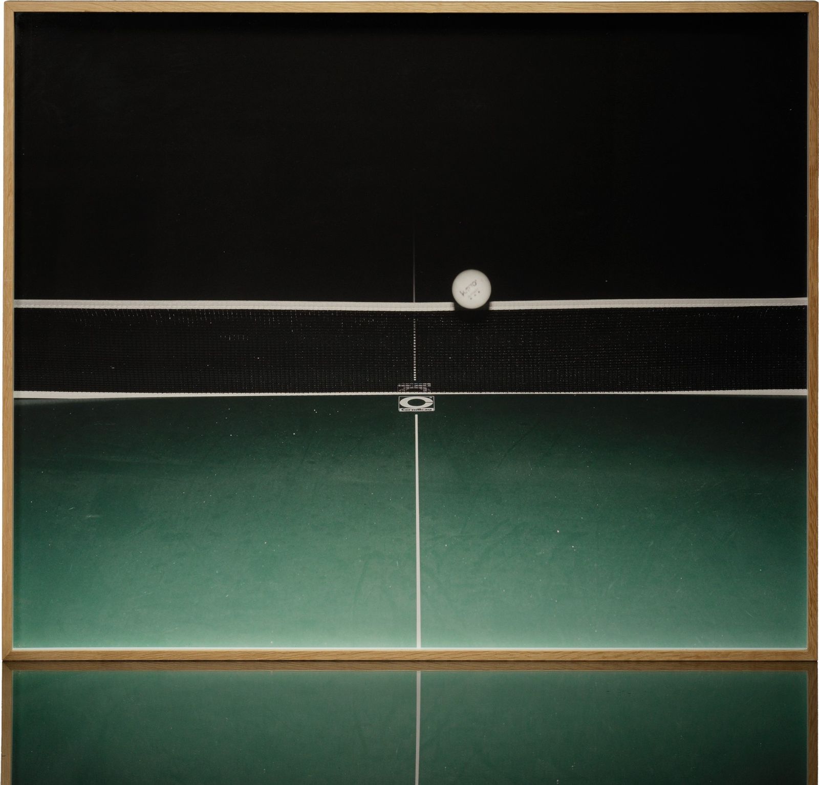 A framed colour print: a table-tennis ball on the top of the net, over a green table, in a light wood frame