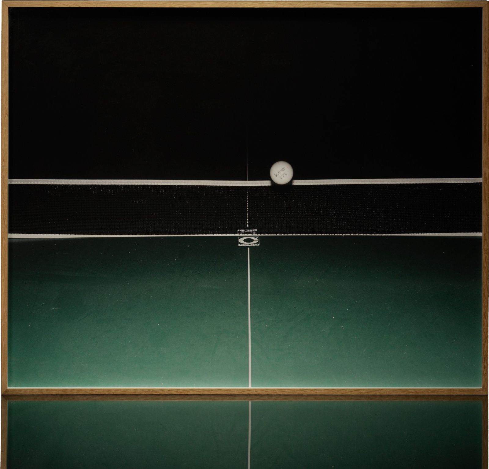 A framed colour print: a table-tennis ball on the top of the net, over a green table, in a light wood frame