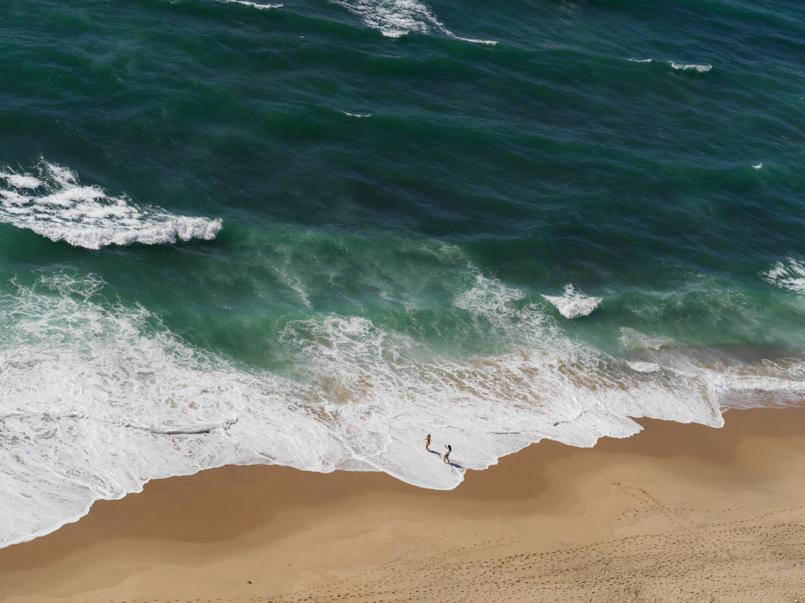 Home Surf breaking on a beach, seen from above, with two figures at the waterline — photograph by Pedro Guimarães